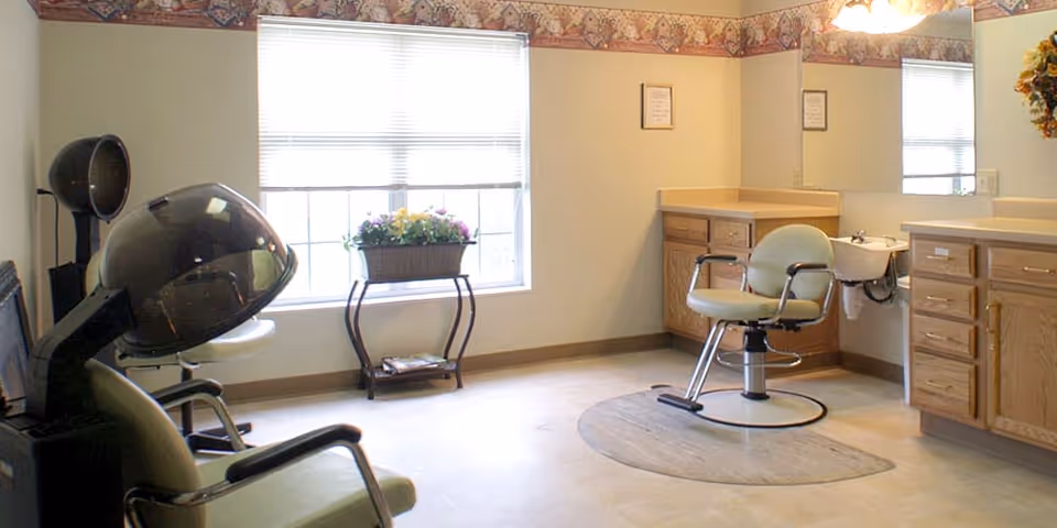 A bright room with two salon chairs, one with a hair dryer hood and the other positioned near a small sink. There is a large window with blinds and a flower arrangement on a small stand beneath it. The walls have a decorative border near the ceiling, and there are wooden cabinets with countertops along one wall.