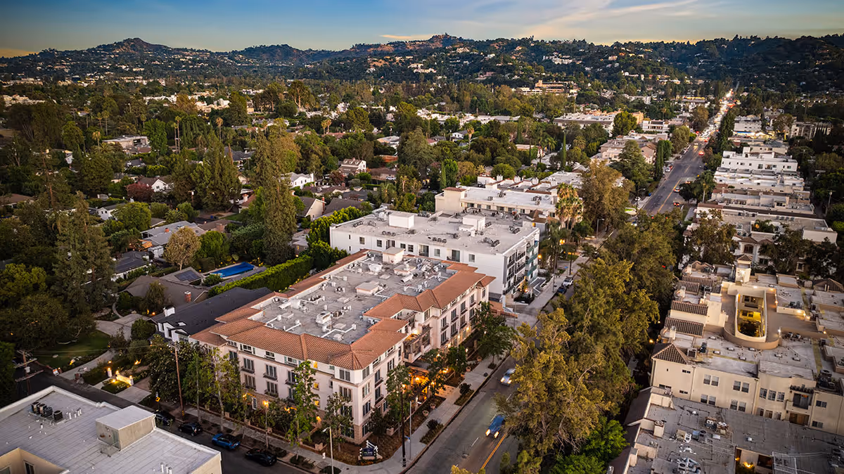 Aerial view of a residential neighborhood at sunset featuring a large multi-story building with a red-tiled roof and white exterior, surrounded by trees and other buildings, with hills visible in the background.