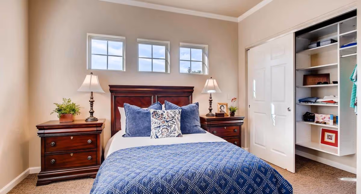 A neatly made bed with a dark wooden headboard and blue patterned bedding in a bedroom. There are two matching wooden nightstands on either side of the bed, each with a lamp and decorative items. Above the bed are three small windows letting in natural light. To the right, there is an open closet with sliding white doors and shelves containing folded clothes and framed pictures.