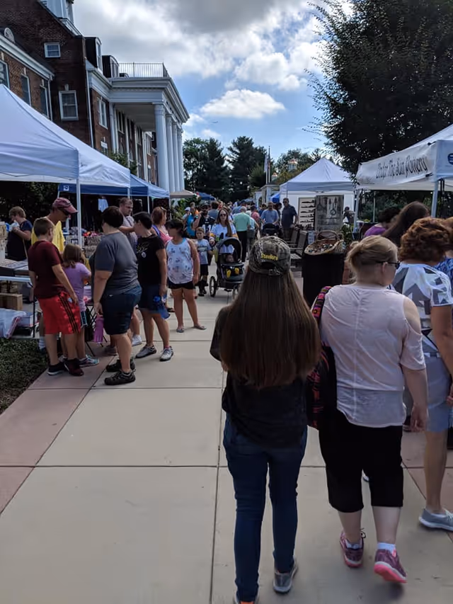 Outdoor scene of a crowded sidewalk market or fair with people walking and browsing stalls set up under white tents next to a large brick building with white columns. Trees and a partly cloudy sky are visible in the background.