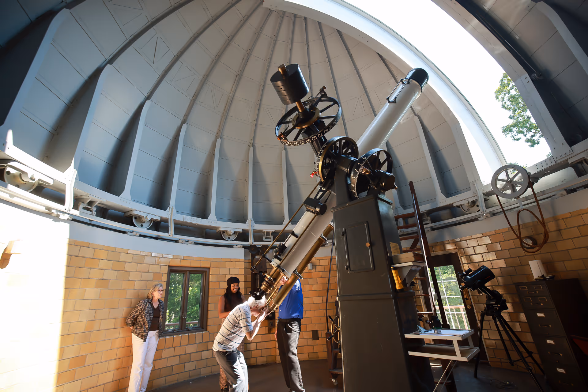 Inside a large observatory dome with a tall telescope in the center. Four people are present: one person is looking through the telescope, another is standing nearby, and two others are observing. The dome has a curved ceiling with a large opening to the sky, and the walls are lined with light-colored bricks and windows.