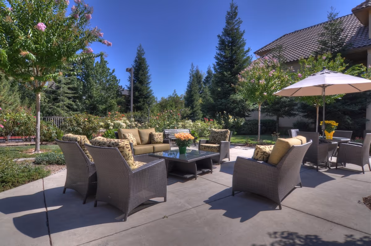 Outdoor patio area with wicker furniture including cushioned chairs and sofas arranged around a glass coffee table. There are trees, shrubs, and flowers surrounding the patio, with a clear blue sky overhead. An umbrella shades a nearby table with chairs.