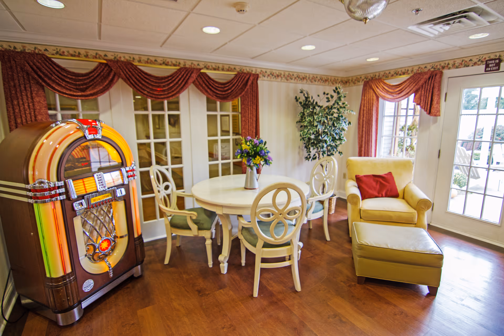 A cozy interior room featuring a vintage jukebox on the left, a round white table with four chairs around it, a vase with colorful flowers on the table, a yellow armchair with a red pillow, and a matching ottoman near a glass door leading outside. The room has wooden flooring, floral wallpaper border, red drapes, and a potted plant in the corner.