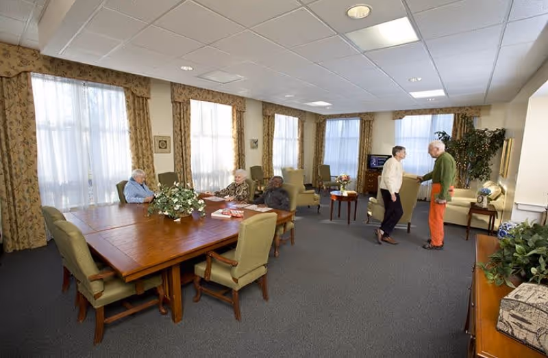 A spacious common room in an assisted living facility with large windows covered by patterned curtains. Three elderly individuals are seated around a wooden table with green upholstered chairs, while two other elderly people are standing and conversing near a seating area with armchairs and a small table. The room is well-lit with ceiling lights and decorated with plants and framed pictures.
