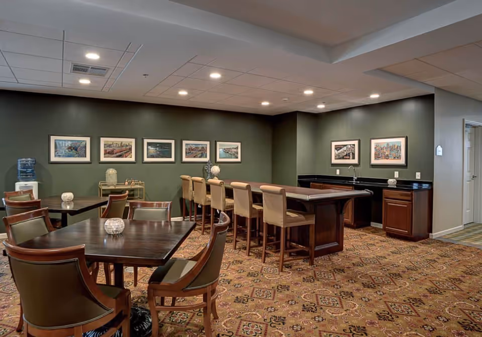 A communal dining and bar area in a senior living facility with dark wooden tables and chairs, a long bar counter with six beige cushioned bar stools, green walls adorned with framed artwork, a water cooler in the corner, and a patterned carpeted floor.