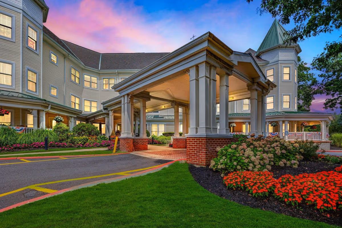 Exterior view of a senior living facility building at sunset with a covered entrance supported by columns, well-maintained landscaping with colorful flowers and green grass, and a multi-story structure with many windows.