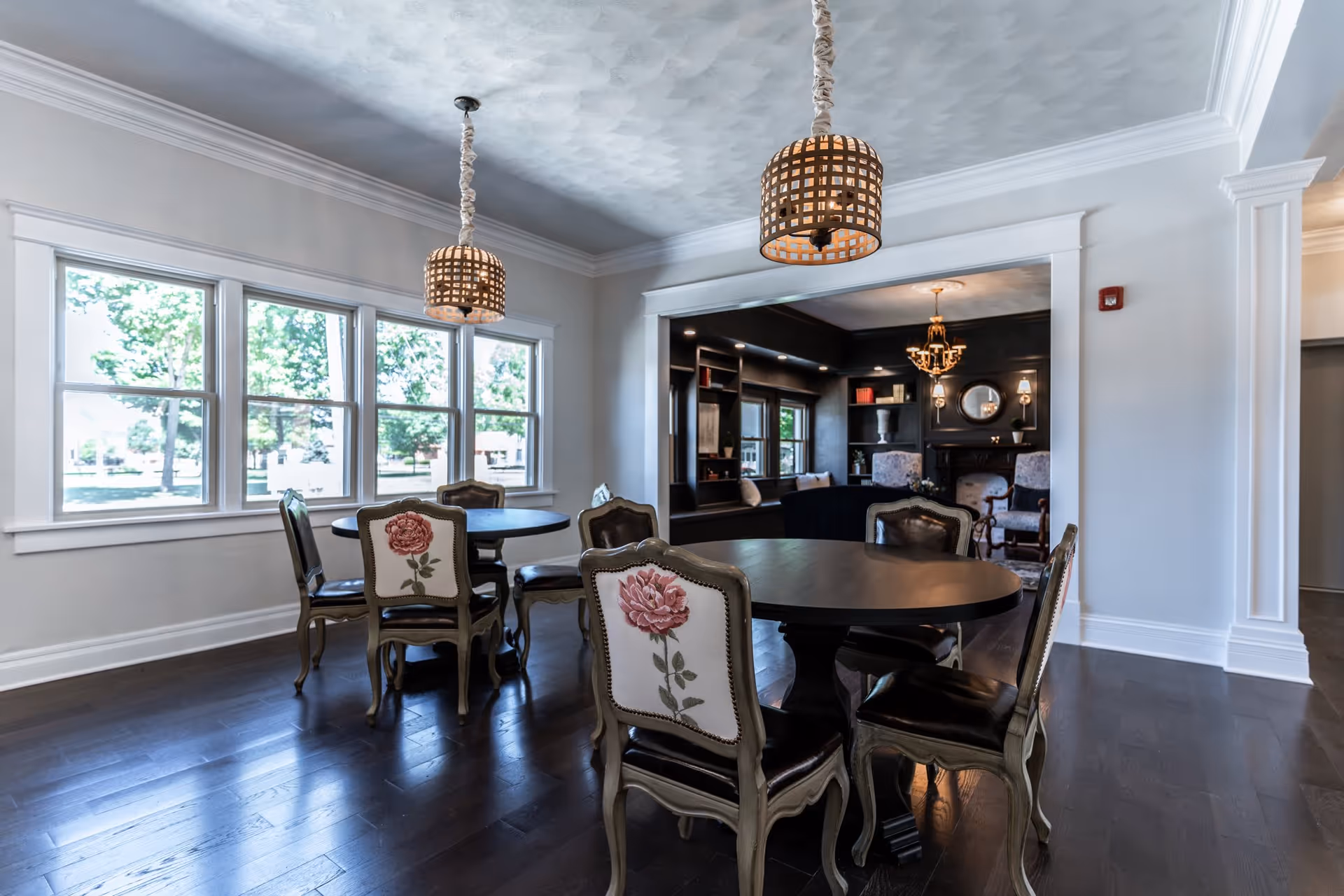 A bright dining area with two round dark wood tables surrounded by chairs with floral upholstery on the backrests. Large windows on one side let in natural light, and two pendant lights with woven shades hang from the ceiling. In the background, there is a cozy living room area with dark walls, built-in shelves, and a chandelier.