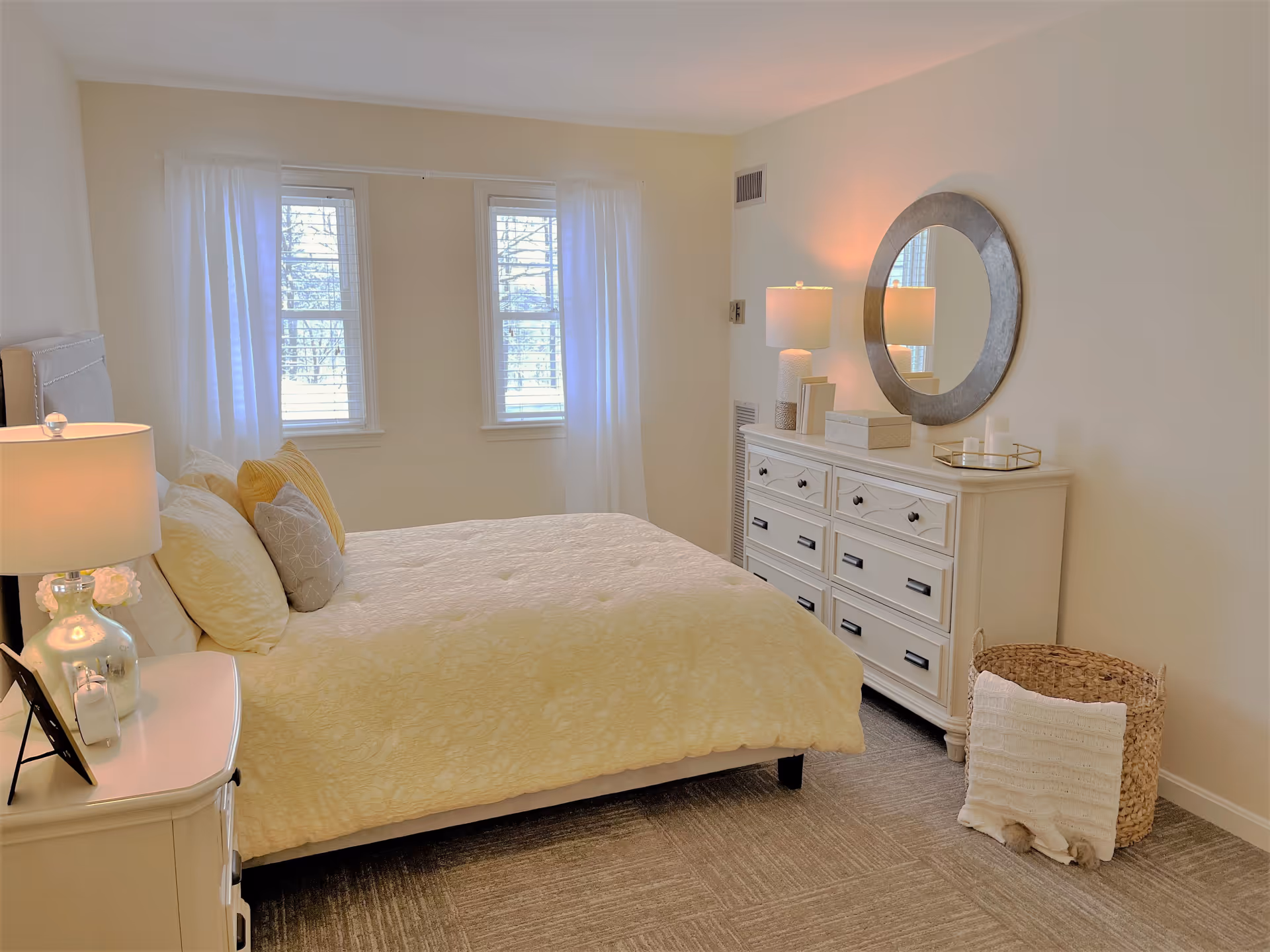 A bright bedroom with a large bed covered in a light yellow comforter and multiple pillows. There are two windows with white curtains letting in natural light. On the right side of the room, there is a white dresser with a round mirror above it, a lamp, and decorative items. A woven basket with a white blanket is placed on the floor next to the dresser. The room has light-colored walls and carpeted flooring.