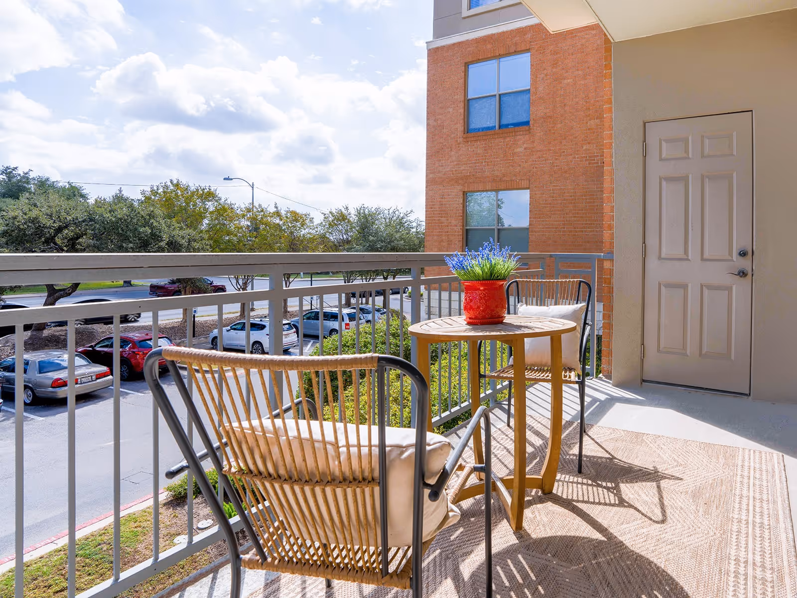 A balcony with two wicker chairs with cushions and a small round wooden table with a red pot of purple flowers. The balcony overlooks a parking lot with several cars and trees, and part of a brick building is visible.