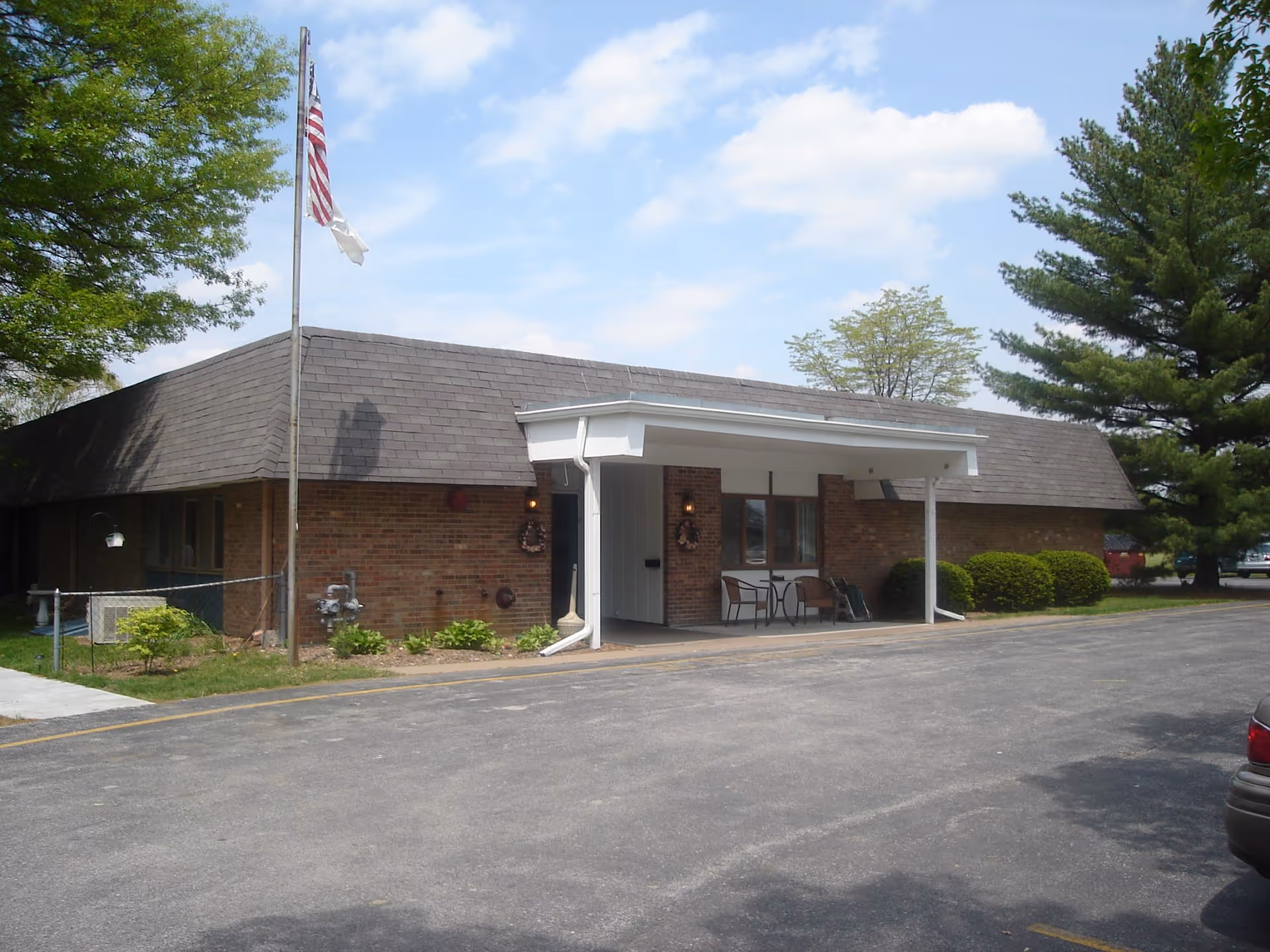 Exterior view of a single-story brick building with a covered entrance, two chairs and a small table on the porch, an American flag on a flagpole, surrounded by trees and bushes under a partly cloudy sky.