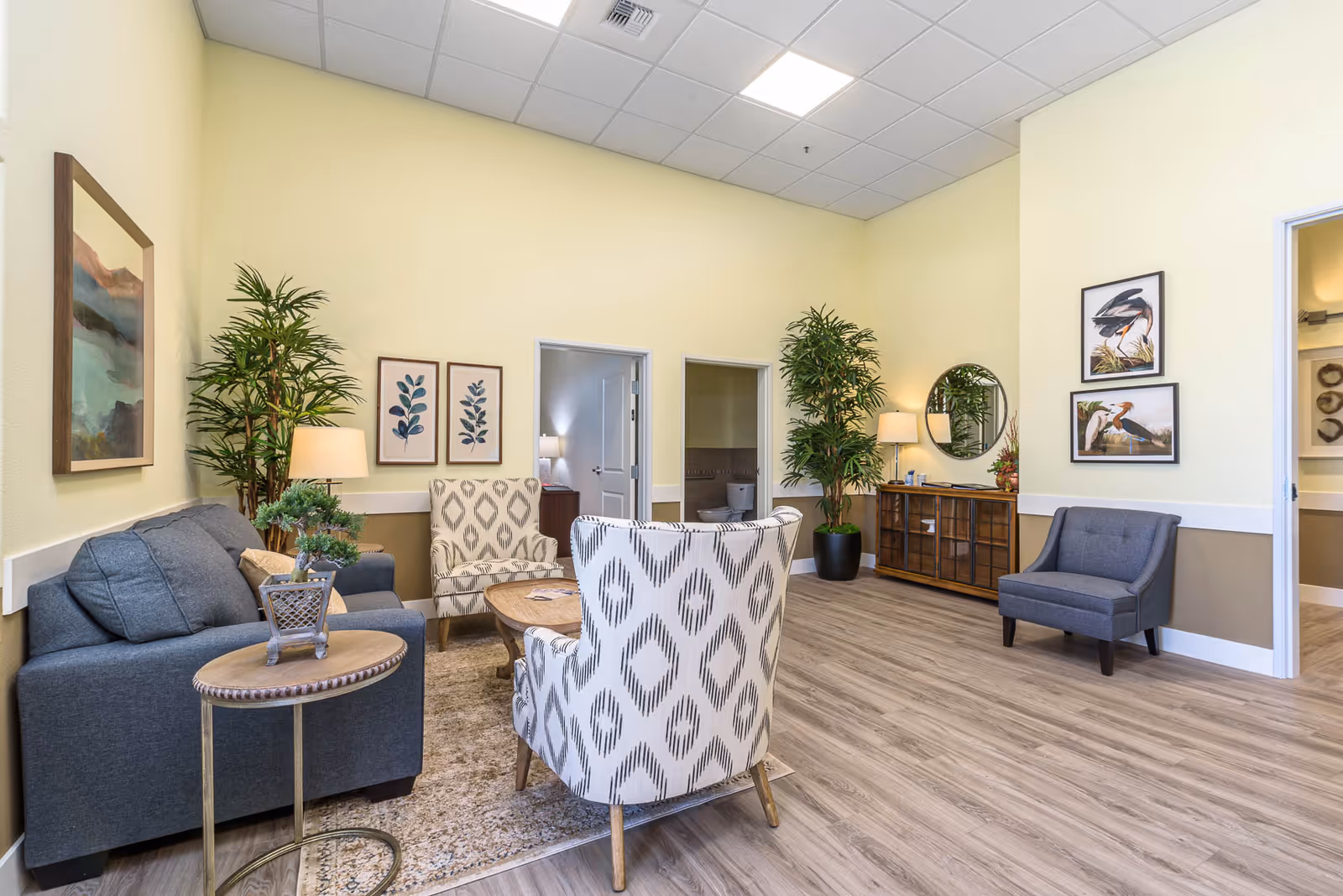 A bright and spacious living room area with light yellow walls and wood flooring. The room features a gray sofa, two patterned armchairs, a round wooden coffee table, and a small side table with a plant. There are two large potted plants, framed botanical and bird artwork on the walls, a wooden cabinet with a round mirror above it, and two open doorways leading to other rooms.