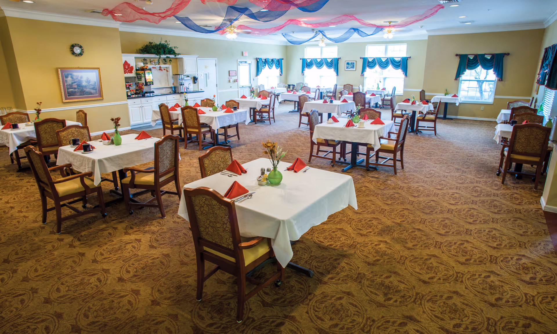 A spacious dining room with multiple tables covered in white tablecloths, each set with red folded napkins, utensils, and small green vases with flowers. The room has patterned carpet, beige walls, windows with teal curtains, and ceiling fans with lights. Red and blue decorative fabric is draped across the ceiling.