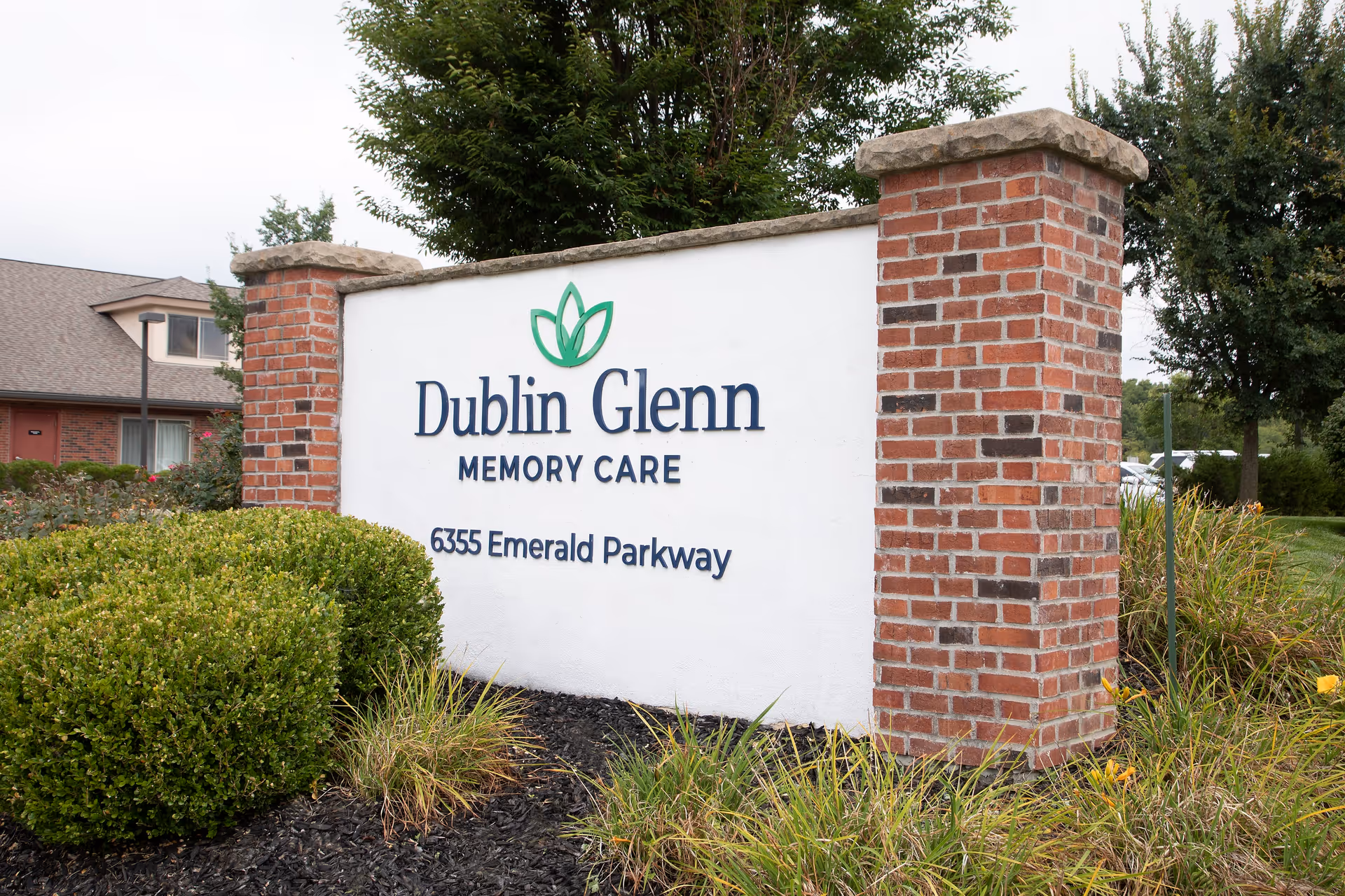 Outdoor sign for Dublin Glenn Memory Care at 6355 Emerald Parkway, surrounded by bushes and landscaping with a brick pillar on the right side.