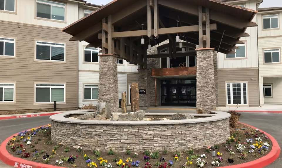 Entrance of Quail Park at Shannon Ranch featuring a covered driveway supported by stone pillars, a circular stone planter with rocks and flowers, and a multi-story building with beige and white siding and multiple windows.