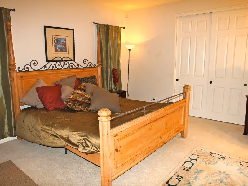 A bedroom with a wooden four-poster bed adorned with multiple pillows in various colors including red, brown, and beige. The bed has a brown comforter. Behind the bed are green curtains and a framed picture on the wall. To the right, there is a floor lamp and white sliding closet doors. The floor is carpeted and a patterned rug is partially visible in the foreground.