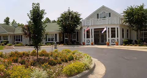 Front exterior view of Brookdale Asheboro facility showing a white building with a green roof, two flags near the entrance, and landscaped garden with trees and flowers in the foreground.