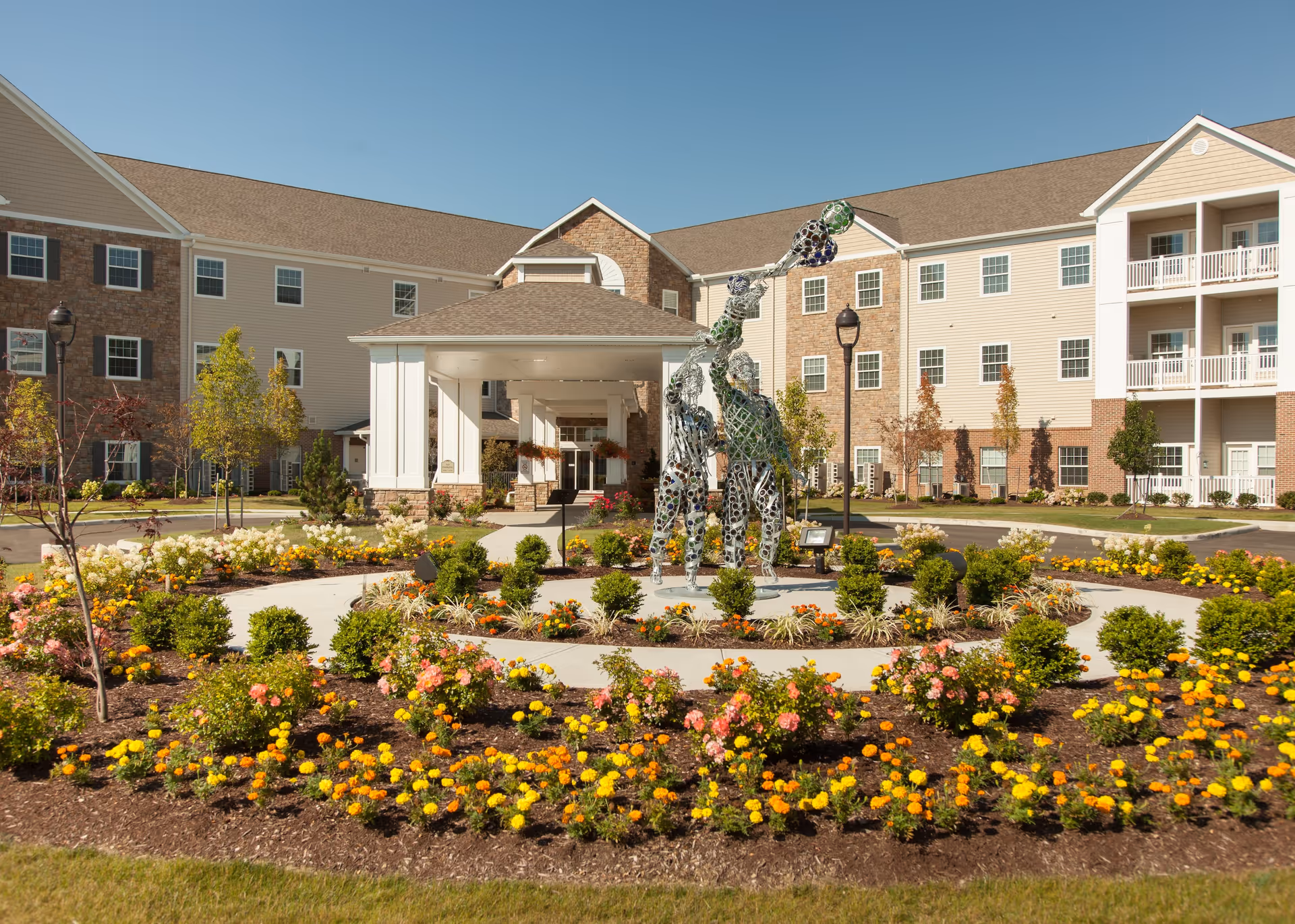 Exterior view of Sanctuary Grande senior living facility with a landscaped circular flower bed featuring a modern metal sculpture of three human figures. The building has multiple stories with beige siding, brick accents, and balconies under a clear blue sky.