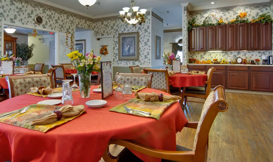 A dining room with round tables covered in red tablecloths, each set with plates, napkins, silverware, and glassware. The chairs have patterned upholstery. There are floral centerpieces on the tables. The room has floral wallpaper, wooden flooring, and wooden cabinets decorated with autumn-themed garlands and flowers.