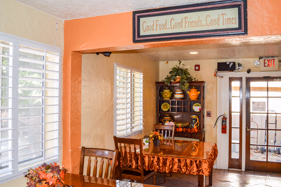 Interior view of a dining area in Rose Garden Court featuring wooden tables with orange tablecloths, wooden chairs, a decorative hutch with colorful pottery, and a sign above the entrance that reads 'Good Food...Good Friends...Good Times'. There are windows with white shutters on the left side and a glass door with an exit sign on the right.