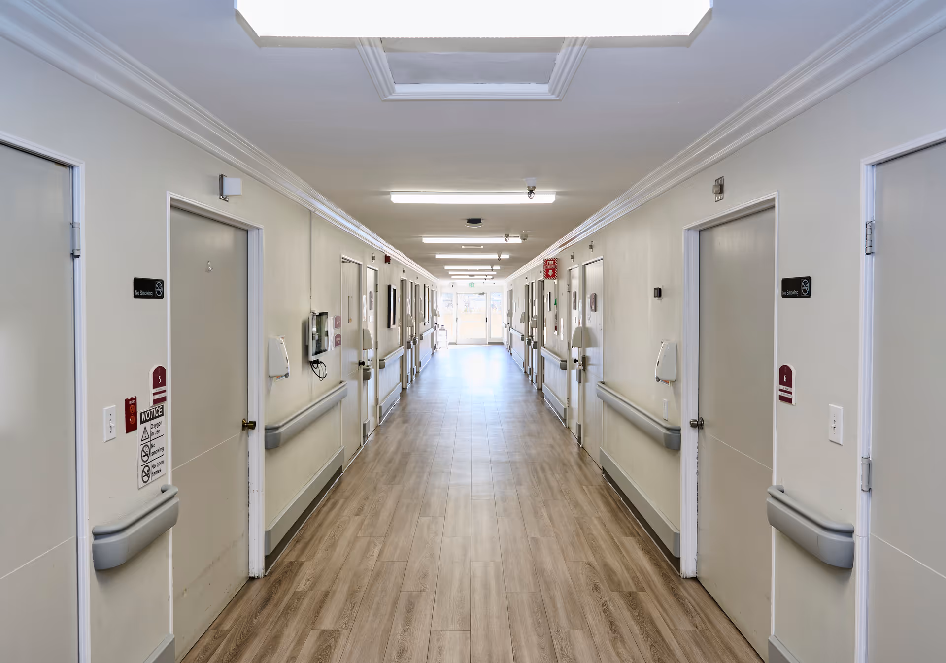 A long, well-lit hallway in a healthcare facility with light-colored walls, wooden flooring, and multiple closed doors on both sides. Handrails run along the walls, and there are ceiling lights and a skylight providing illumination. The hallway leads to a glass door at the far end.