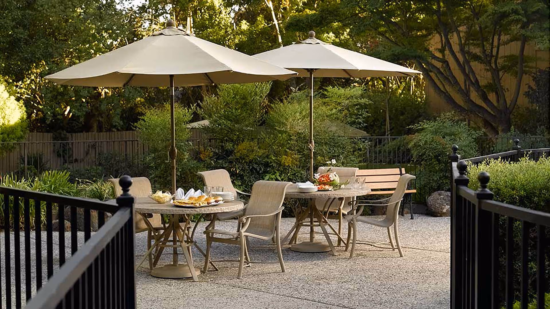 Outdoor patio area with two round tables, each shaded by a large beige umbrella. Each table is surrounded by beige chairs and set with plates of food and napkins. The patio is enclosed by a black metal fence and surrounded by green bushes and trees.