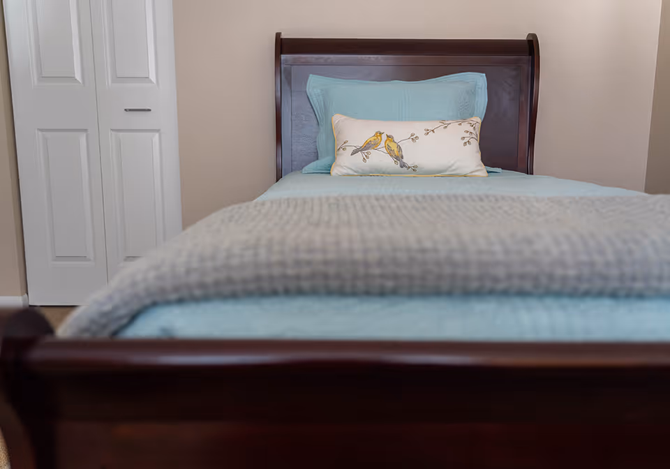 A neatly made single bed with a wooden headboard and footboard. The bed is covered with a light blue quilt and a gray knitted blanket at the foot. There are two pillows, one light blue and one decorative with a bird design. A white closet door is visible on the left side against a beige wall.
