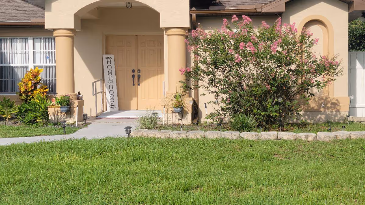 Front entrance of a beige building with double doors, two columns, a small garden with green plants and pink flowers, and a sign leaning against one column that reads 'HOME'.
