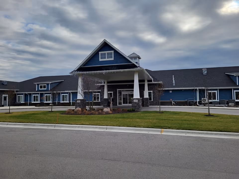 Front exterior view of a single-story building with blue siding and a dark roof, featuring a covered entrance supported by white pillars. The building is surrounded by a well-maintained lawn and a paved driveway under a cloudy sky.
