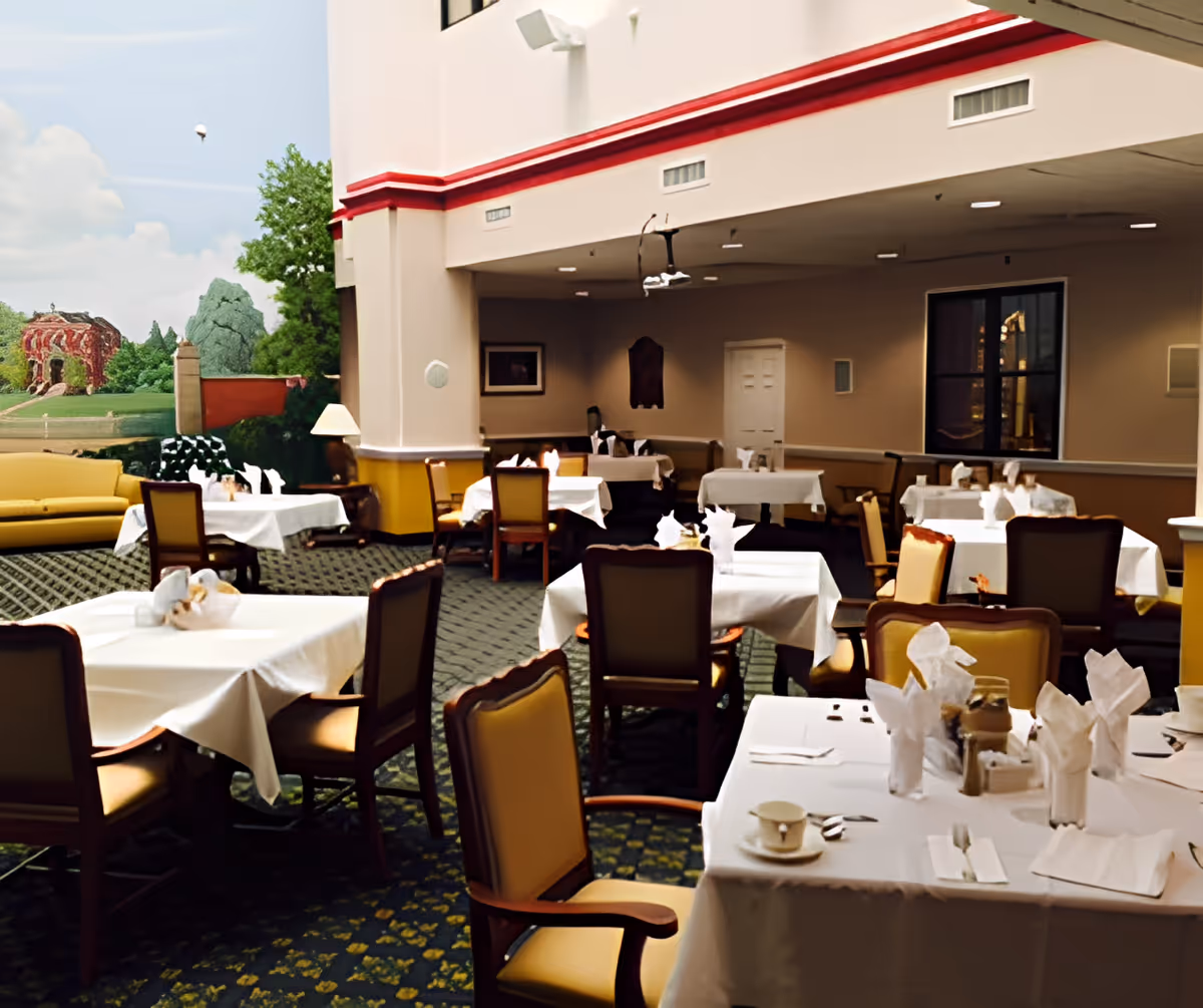 Interior view of a dining room with multiple tables covered in white tablecloths, each set with napkins, cups, and utensils. The room has cushioned chairs around the tables, a patterned carpet, and a mural of a garden scene on one wall. There is a yellow couch and a lamp near the mural, and the ceiling has recessed lighting.