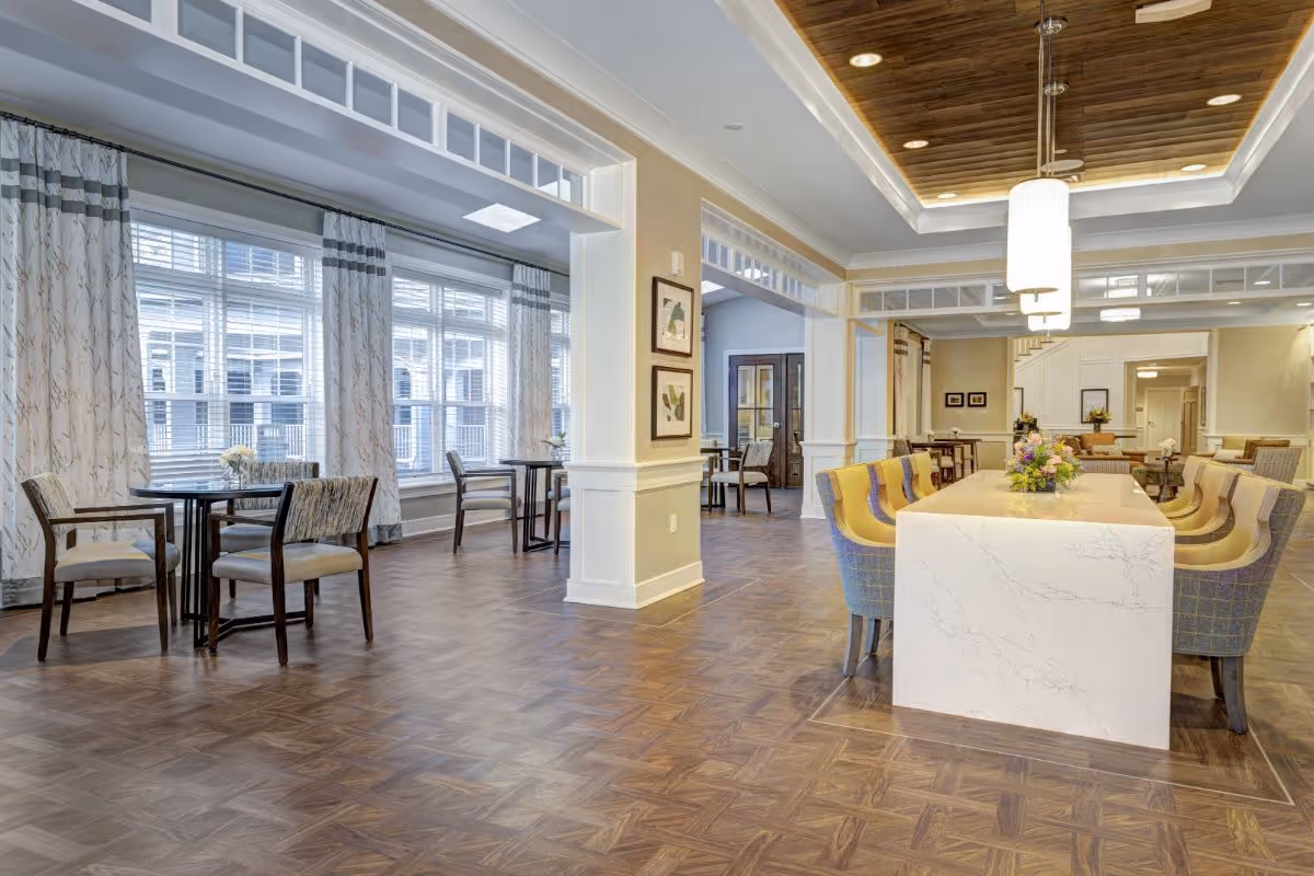 A spacious and well-lit common area in a senior living facility with large windows covered by patterned curtains. The room features a long white marble table with several upholstered chairs around it, smaller round tables with chairs near the windows, wooden flooring, and a ceiling with recessed lighting and hanging pendant lights. The decor includes framed artwork on the walls and flower arrangements on the tables.