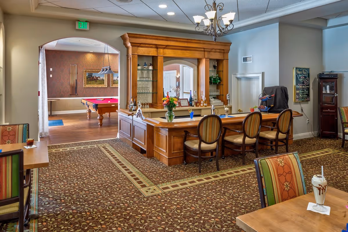 Interior view of a senior living facility lounge area with a wooden bar counter and three chairs. The bar has flowers, drinks, and glassware on display. To the left, there is an arched doorway leading to a room with a pool table and framed artwork on the wall. The room has patterned carpet and a chandelier hanging from the ceiling. A chalkboard on the wall lists happy hour drink specials.