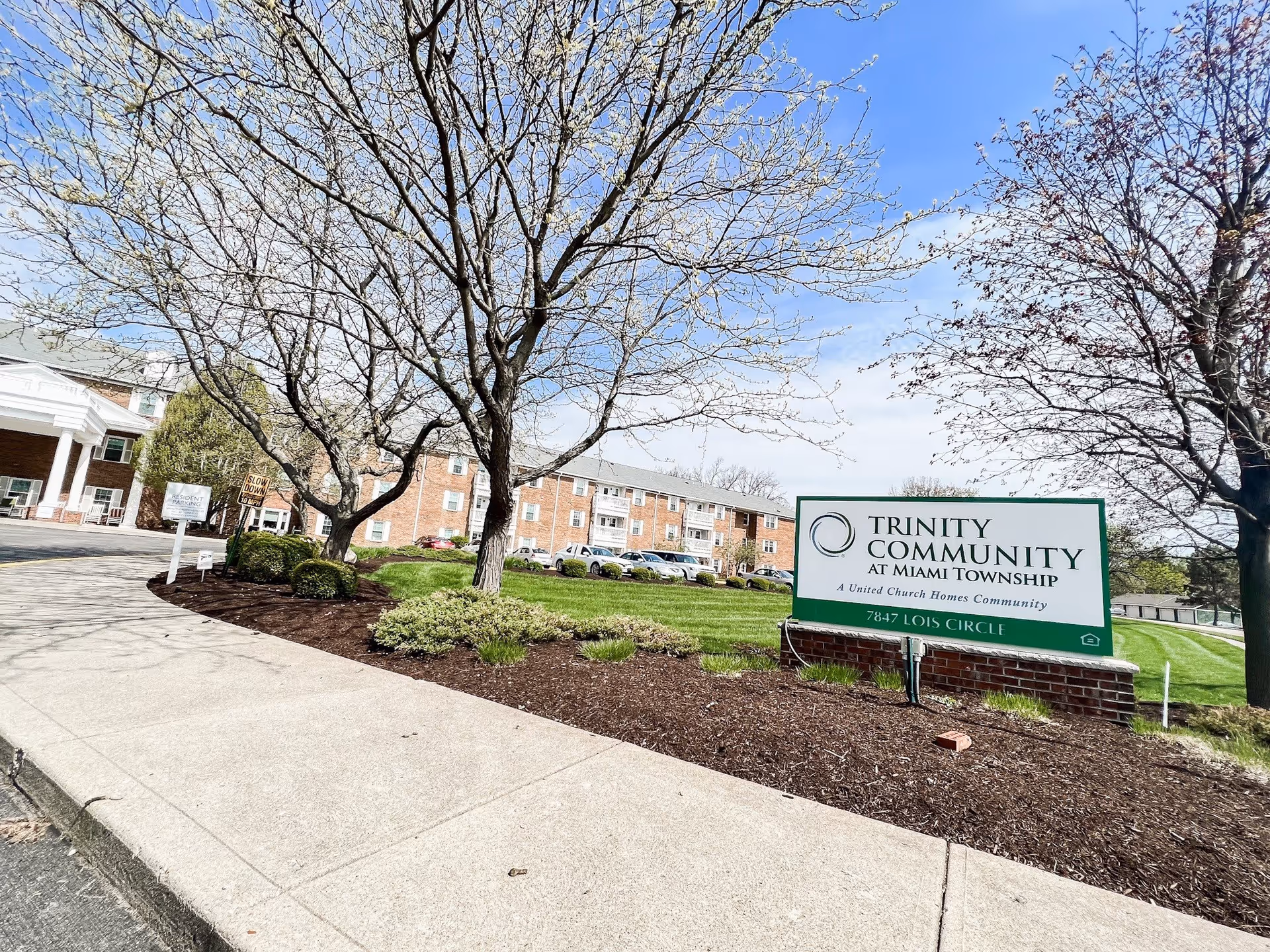 Exterior view of The Trinity Community at Miami Township senior living facility showing a sidewalk, landscaped area with trees and bushes, a large sign with the facility name and address, and a multi-story brick building in the background under a partly cloudy sky.
