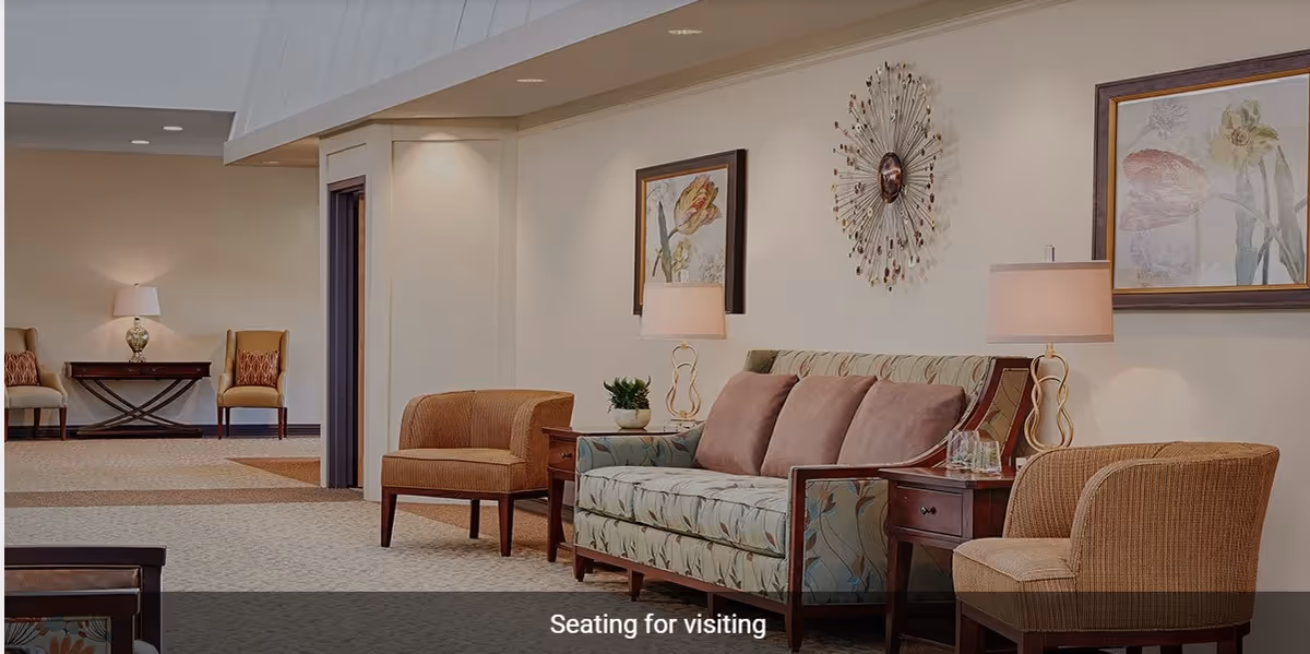 A cozy seating area in a senior living facility with a floral-patterned sofa, two cushioned armchairs, side tables with lamps, and framed floral artwork on the walls.