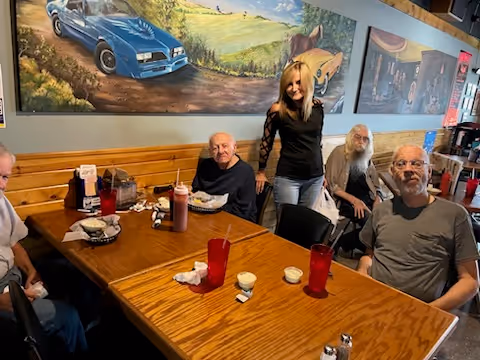 A group of four elderly men and a younger woman gathered around a wooden dining table in a casual dining setting. The table has red plastic cups, condiments, and plates with food. The background features a mural with vintage cars and a countryside scene.