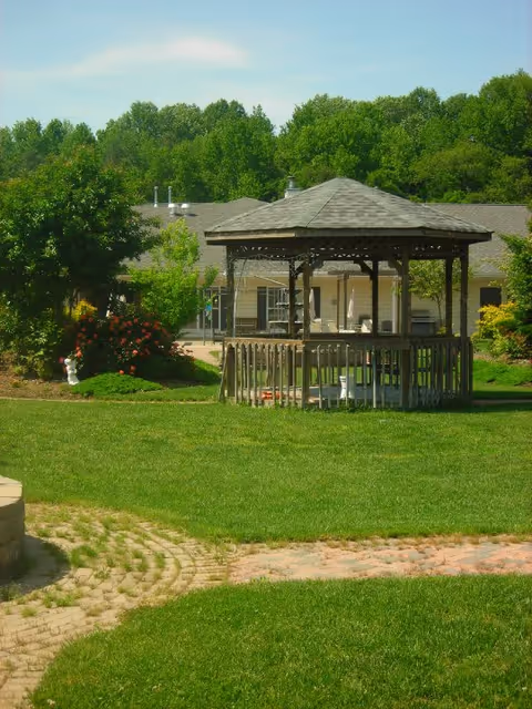 A wooden gazebo with a shingled roof situated on a well-maintained grassy lawn. Behind the gazebo, there is a single-story building partially visible with beige siding and several windows. Surrounding the area are green trees and bushes, and a paved stone pathway curves through the grass in the foreground.
