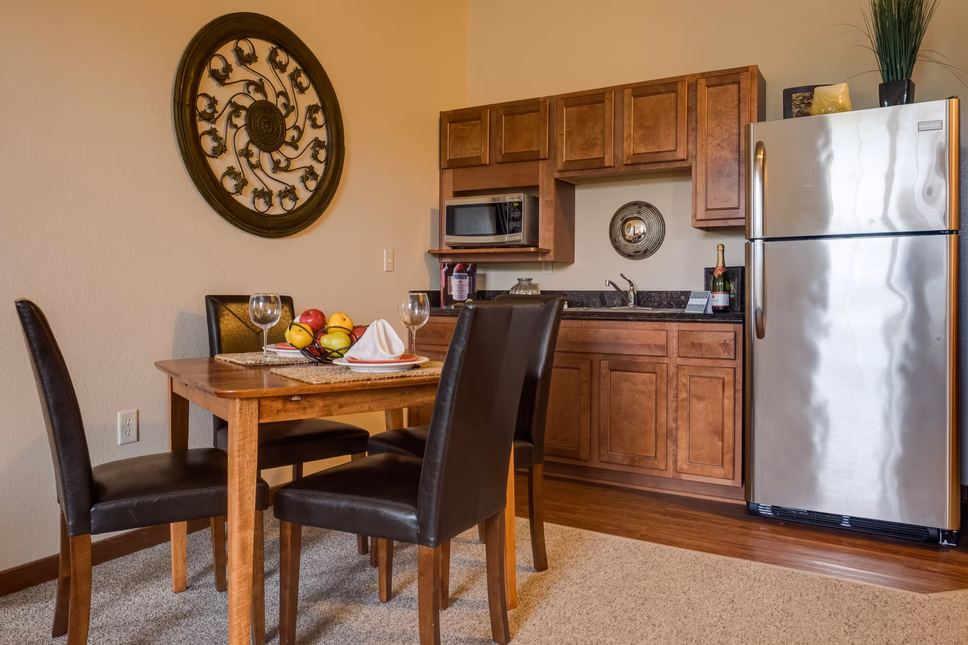 A small kitchen and dining area featuring a wooden table set with four dark brown leather chairs. The table has two place settings with plates, napkins, and wine glasses, and a fruit basket with apples and lemons. The kitchen area includes wooden cabinets, a microwave, a sink, and a stainless steel refrigerator. A decorative round metal wall art is mounted on the wall above the dining table, and a small plant and decorative items are placed on top of the refrigerator.