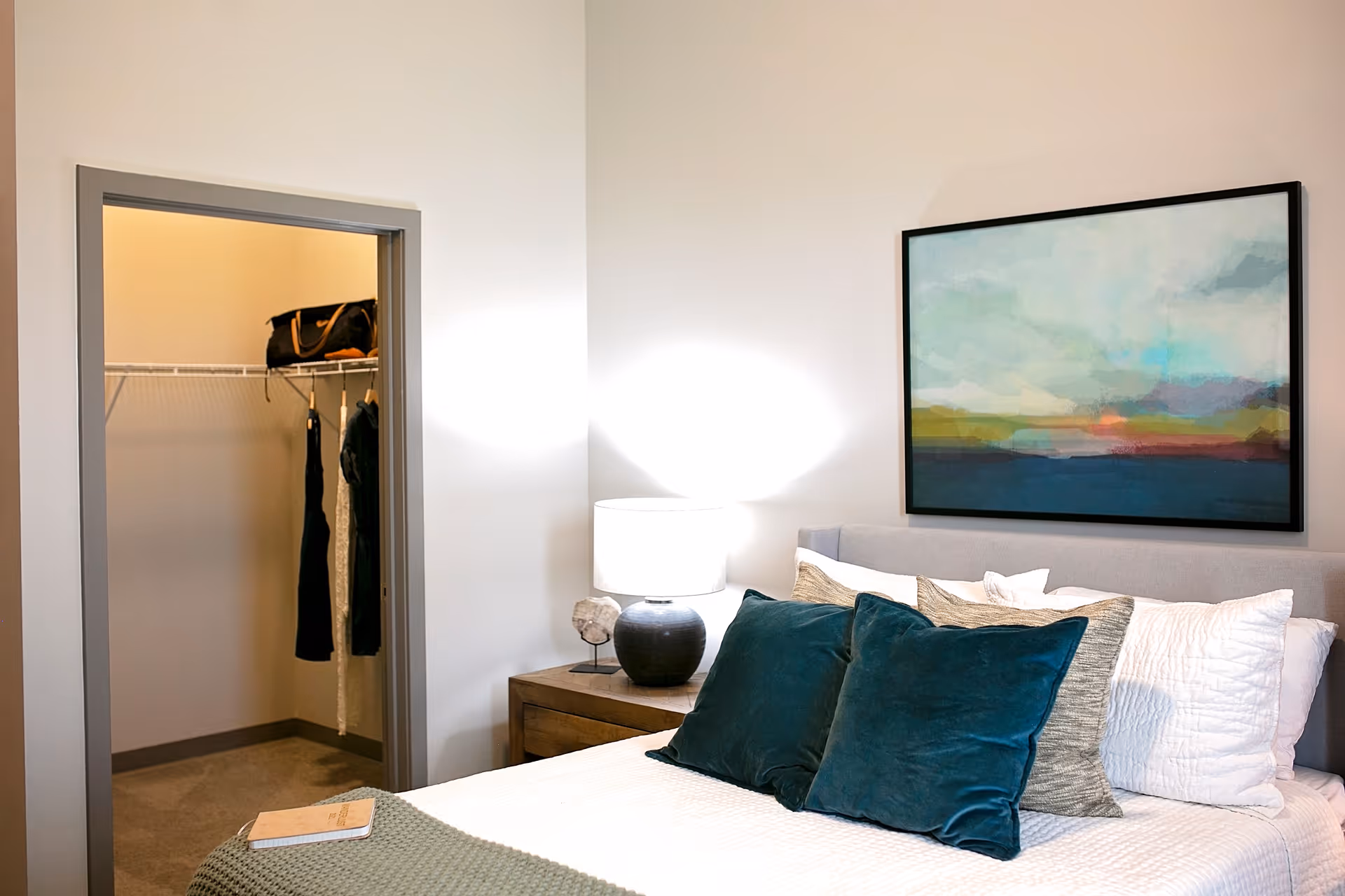 A cozy bedroom with a neatly made bed featuring white and teal pillows, a wooden nightstand with a lamp, and an abstract landscape painting on the wall. An open doorway reveals a walk-in closet with hanging clothes and a bag on the shelf.