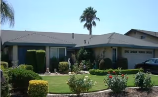 Single-story residential building with a well-maintained front yard featuring green grass, trimmed bushes, flowering plants, and a palm tree under a clear blue sky.