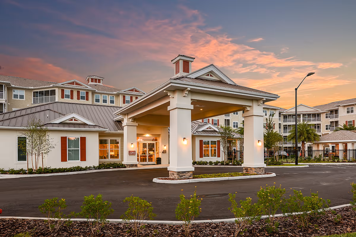 Exterior view of Album Monterey Pointe 55+ Active Adult Apartment Homes at sunset, showing the main entrance with a covered driveway, well-maintained landscaping, and multi-story residential buildings in the background.