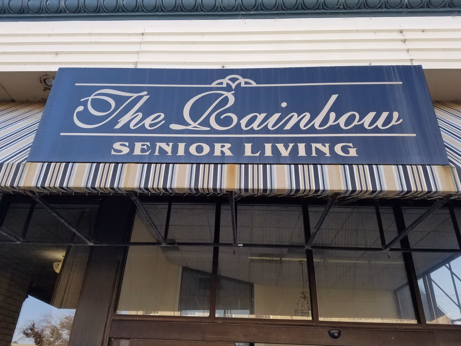 A blue and white striped awning with a sign that reads 'The Rainbow Senior Living' in elegant white script and block letters above the entrance of a building.