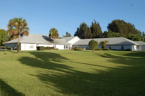 Single-story white building with a gray roof surrounded by a large, well-maintained grassy lawn and several palm trees under a clear blue sky.