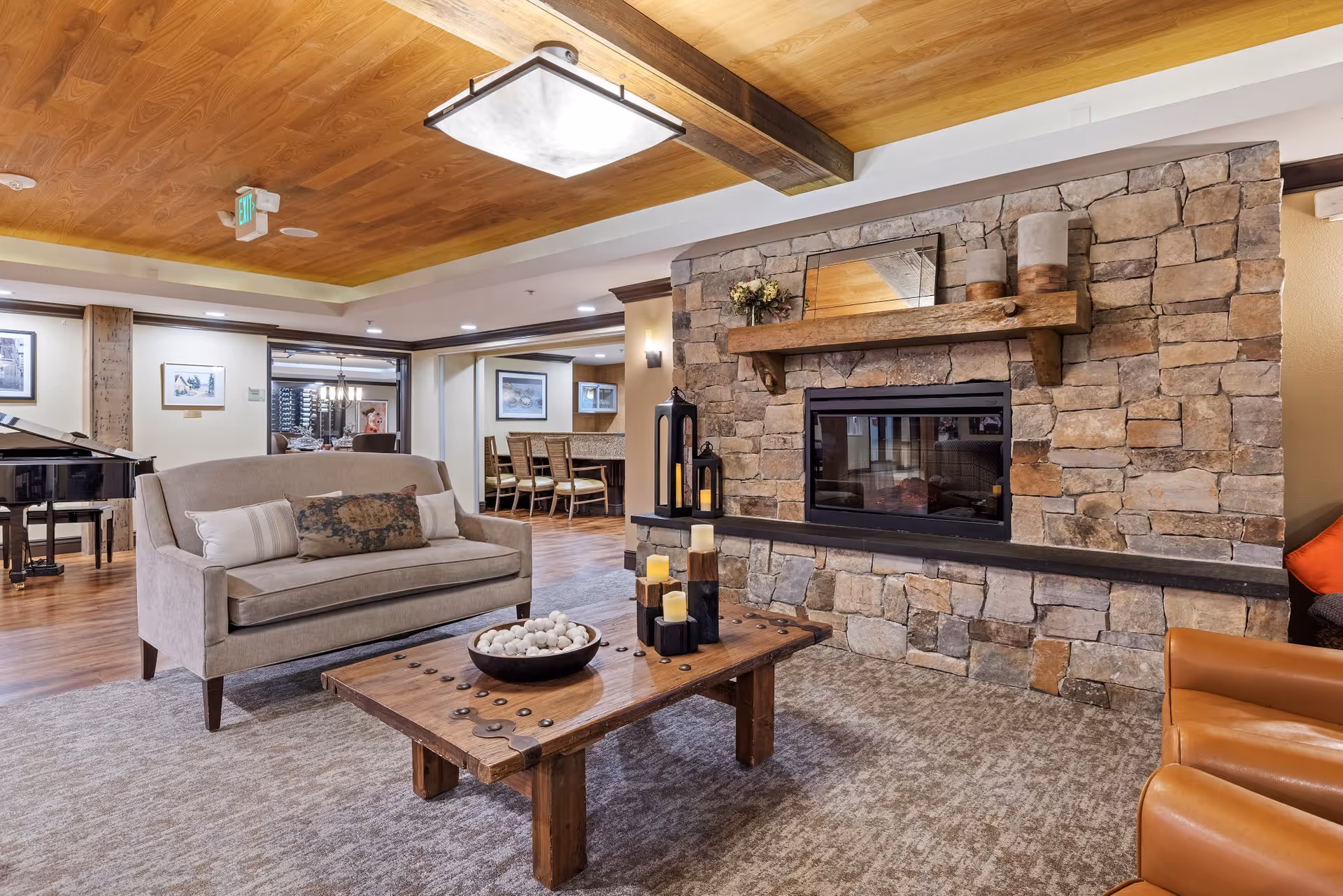 Cozy living room area with a stone fireplace, wooden mantel decorated with candles and a small flower arrangement. A beige loveseat with decorative pillows faces a rustic wooden coffee table adorned with candles and a bowl of decorative balls. The room features a wooden ceiling with exposed beams, a large ceiling light, and hardwood flooring. In the background, there is a dining area with a table and chairs, and a black grand piano is visible to the left.