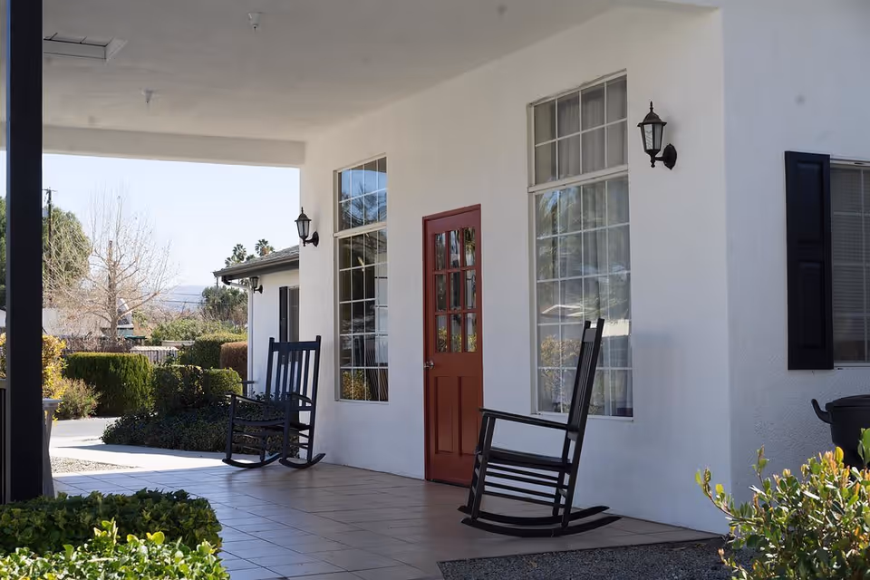 A covered outdoor porch area with two black rocking chairs placed on tiled flooring. The porch is attached to a white building with large windows and a red door. There are wall-mounted lantern-style lights on either side of the door. Shrubs and trees are visible in the background under a clear sky.