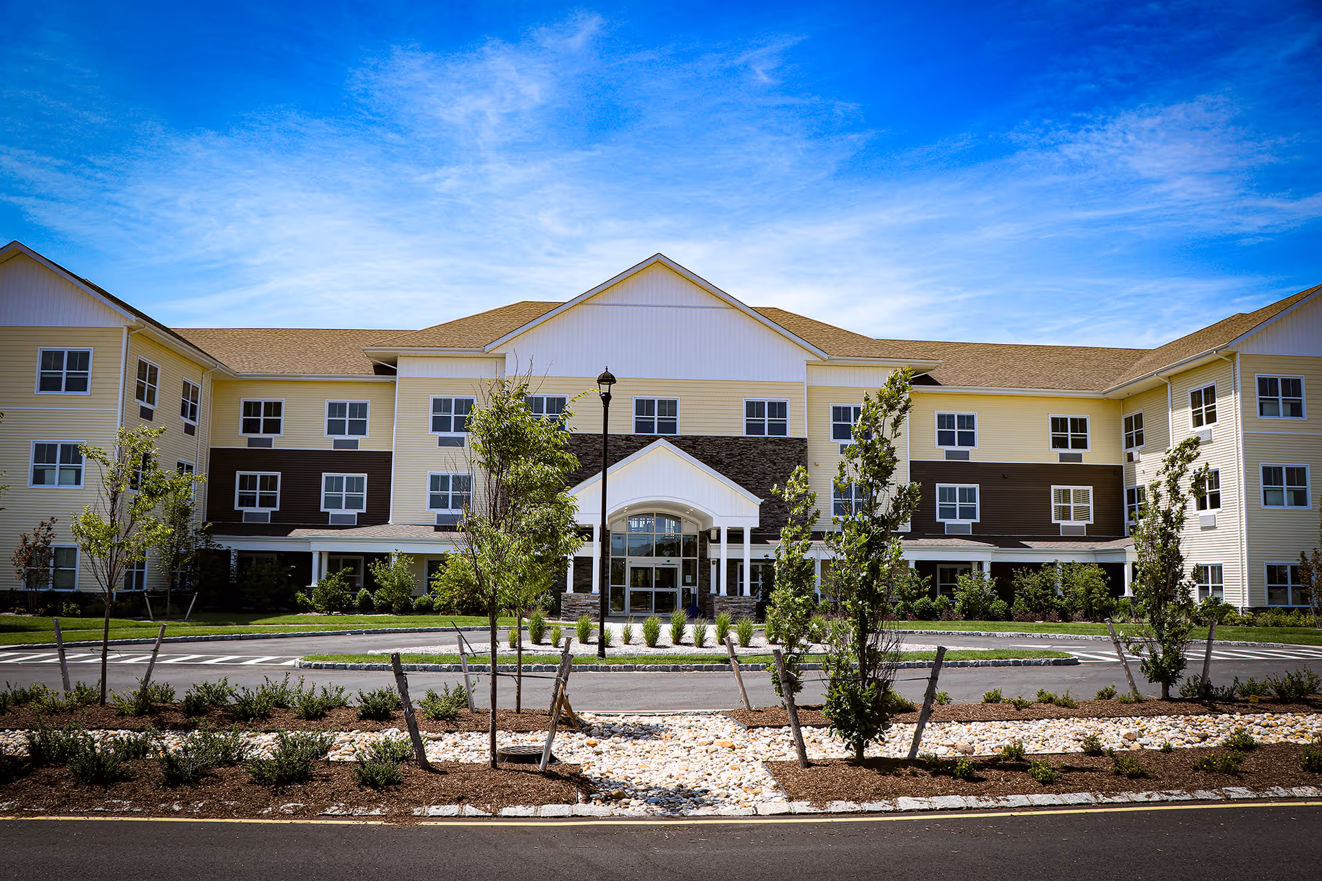 Front exterior view of a three-story senior living facility building with a beige and brown facade, multiple windows, a central entrance with a covered porch, landscaped garden with young trees and shrubs, and a clear blue sky above.
