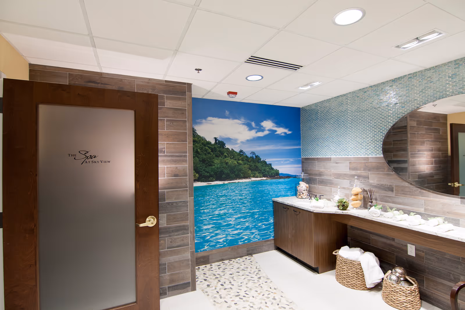 Interior view of a spa room with a wooden door labeled 'The Spa at Sky View'. The room features a large wall mural of a tropical beach with blue water and green trees. There is a long countertop with rolled white towels, decorative jars, and small plants. The wall above the counter has blue mosaic tiles and a large oval mirror. Two wicker baskets with towels are placed under the counter.