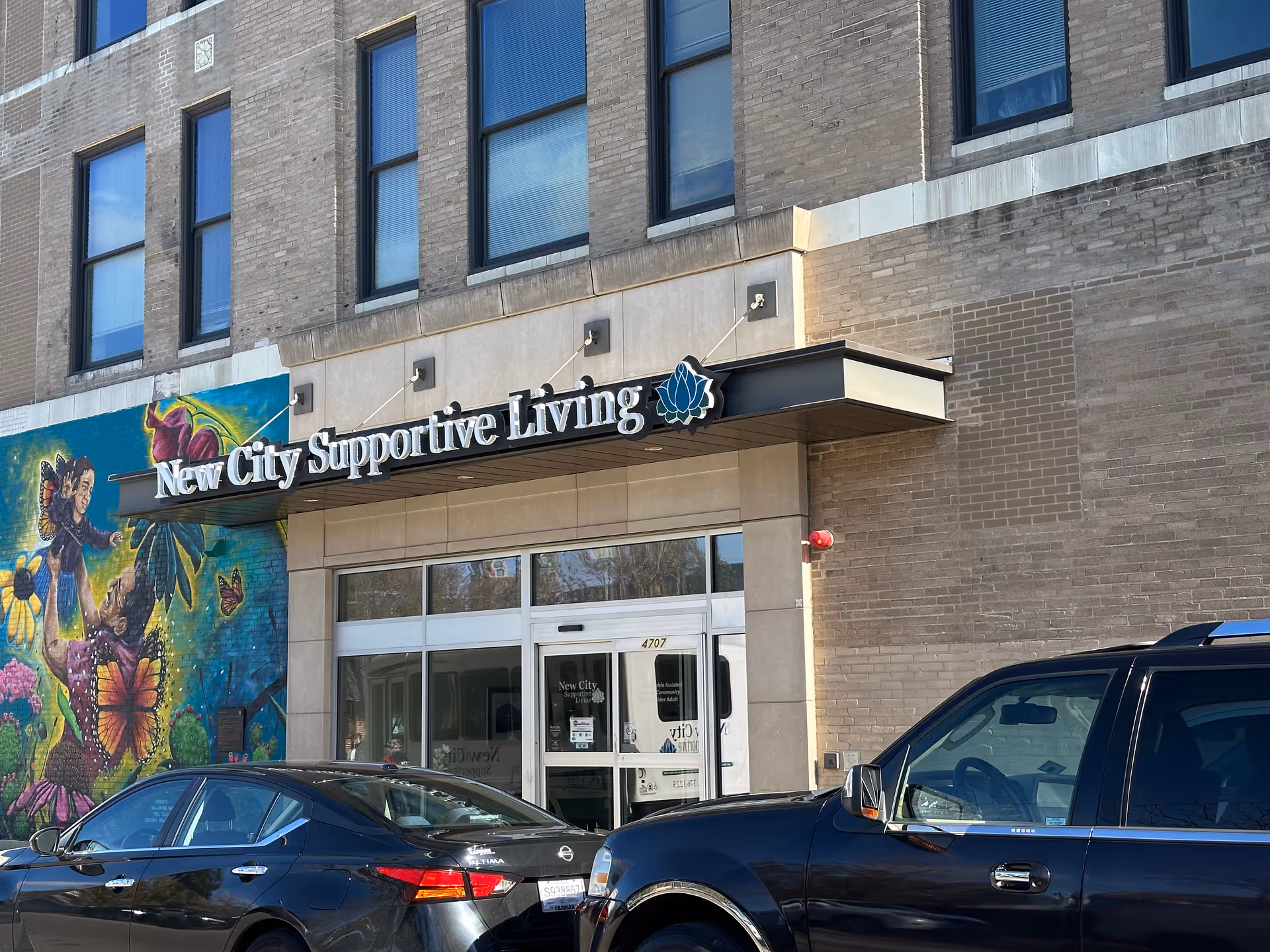 Exterior view of the New City Supportive Living facility showing the building entrance with a sign above the door. There is a colorful mural on the left side of the entrance depicting a person holding a child surrounded by butterflies and flowers. Two cars are parked in front of the building.