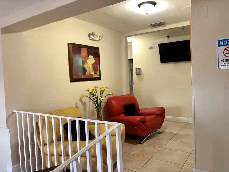 A small seating area inside a retirement home with a yellow armchair and a red armchair, a vase with yellow flowers between them, a framed painting of vases on the wall, a wall-mounted TV, and a no smoking sign on the wall.