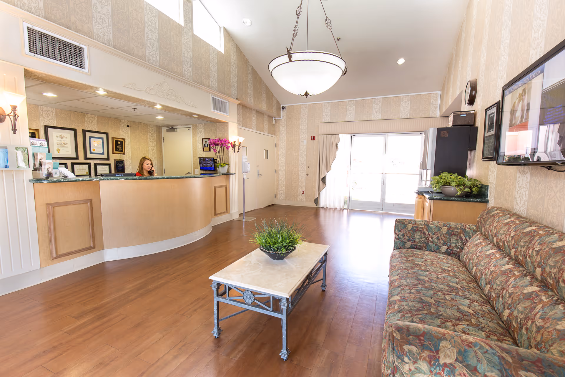 Reception area of Windsor Terrace Healthcare Center with a curved wooden front desk where a woman is seated. The room has wooden flooring, a floral patterned couch, a coffee table with a plant, framed certificates on the wall behind the desk, a large hanging light fixture, and a glass door letting in natural light.