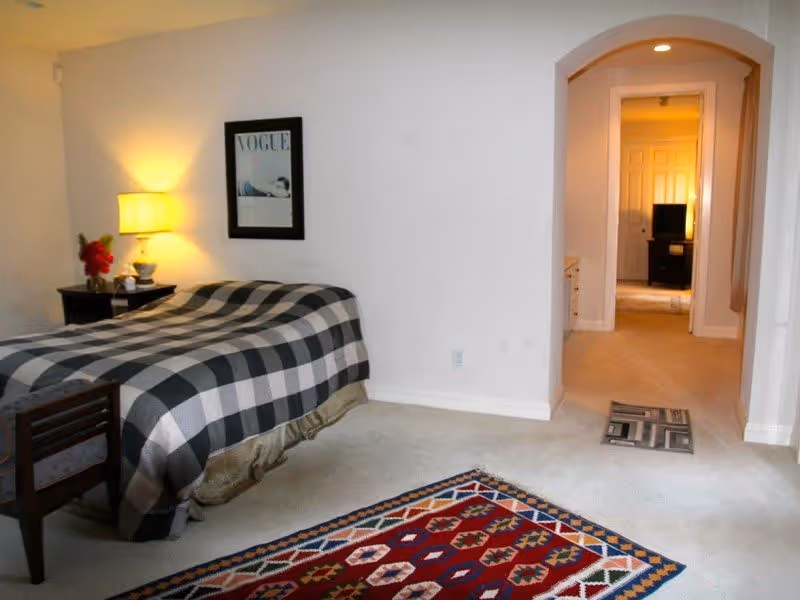 A bedroom with a bed covered in a black and white checkered blanket, a wooden chair, a nightstand with a lamp and flowers, a framed Vogue magazine cover on the wall, and a colorful patterned rug on the carpeted floor. There is an arched doorway leading to another room with a TV and closed doors.