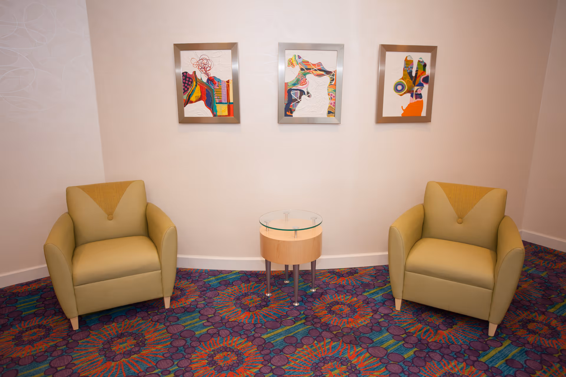 Two light green armchairs with a small round wooden table with a glass top between them, set against a white wall with three colorful abstract paintings hanging above. The floor is covered with a vibrant patterned carpet in purple, blue, orange, and green tones.